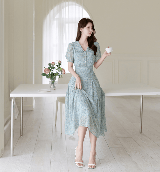 Woman in romantic floral chiffon dress with frill collar and waist ribbon, sitting gracefully with a cup in elegant, airy room.