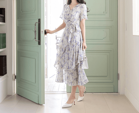 Woman wearing asymmetrical floral chiffon dress with purple blossoms, puffed sleeves, and V-neck, standing by a green door.