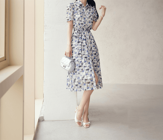 Woman wears breezy chiffon shirt dress with blue abstract pattern and belt, standing in bright room with stylish purse and heels.