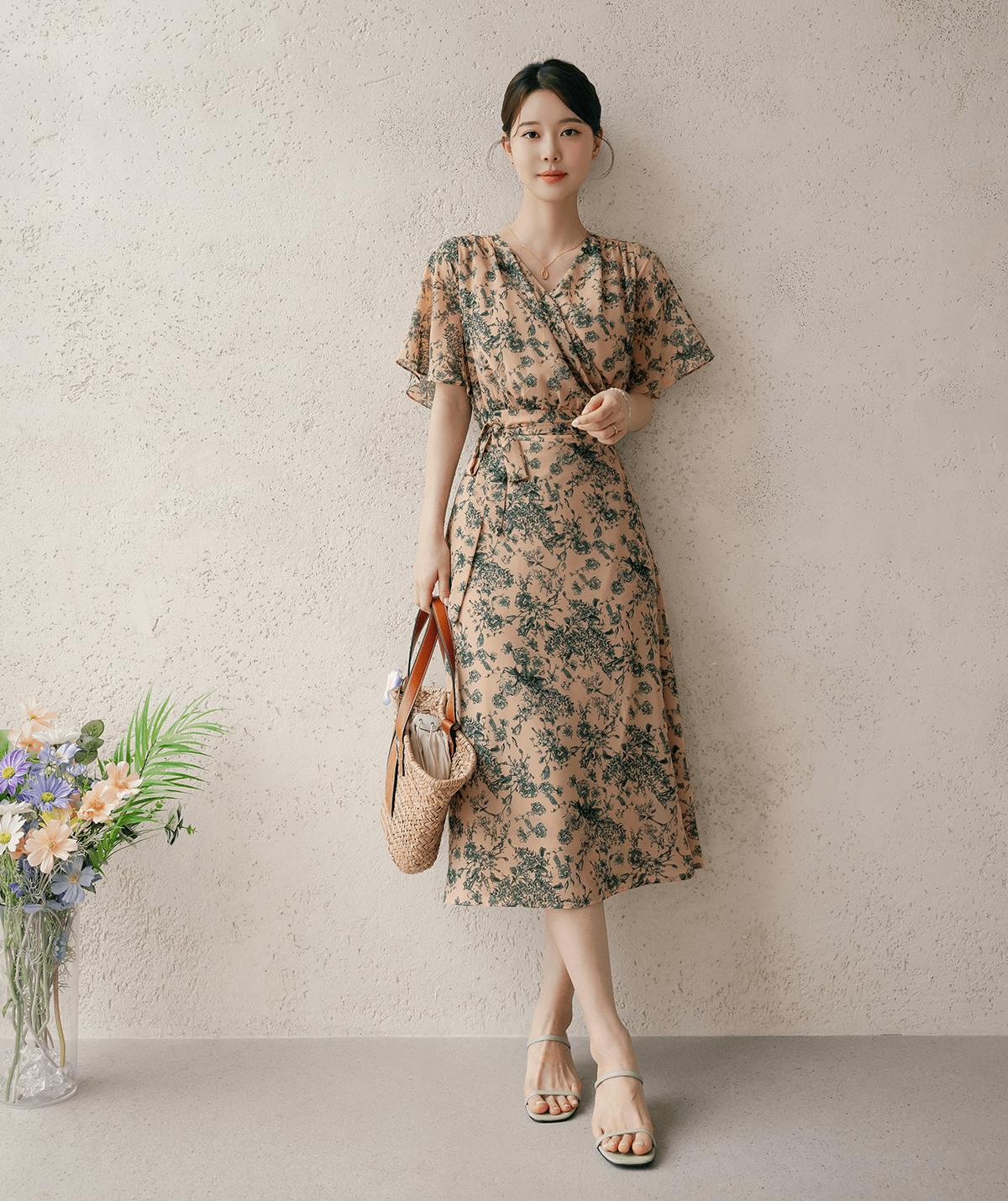 Woman wearing a goddess-inspired wrap dress with graceful wing sleeves, holding a straw bag, standing against a textured wall.