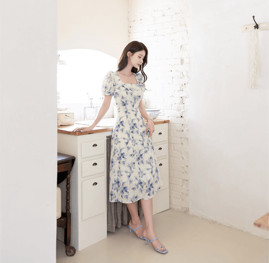 Elegant chiffon dress with blue-violet abstract print and square ruffle neckline, modeled in a bright, minimalist room.