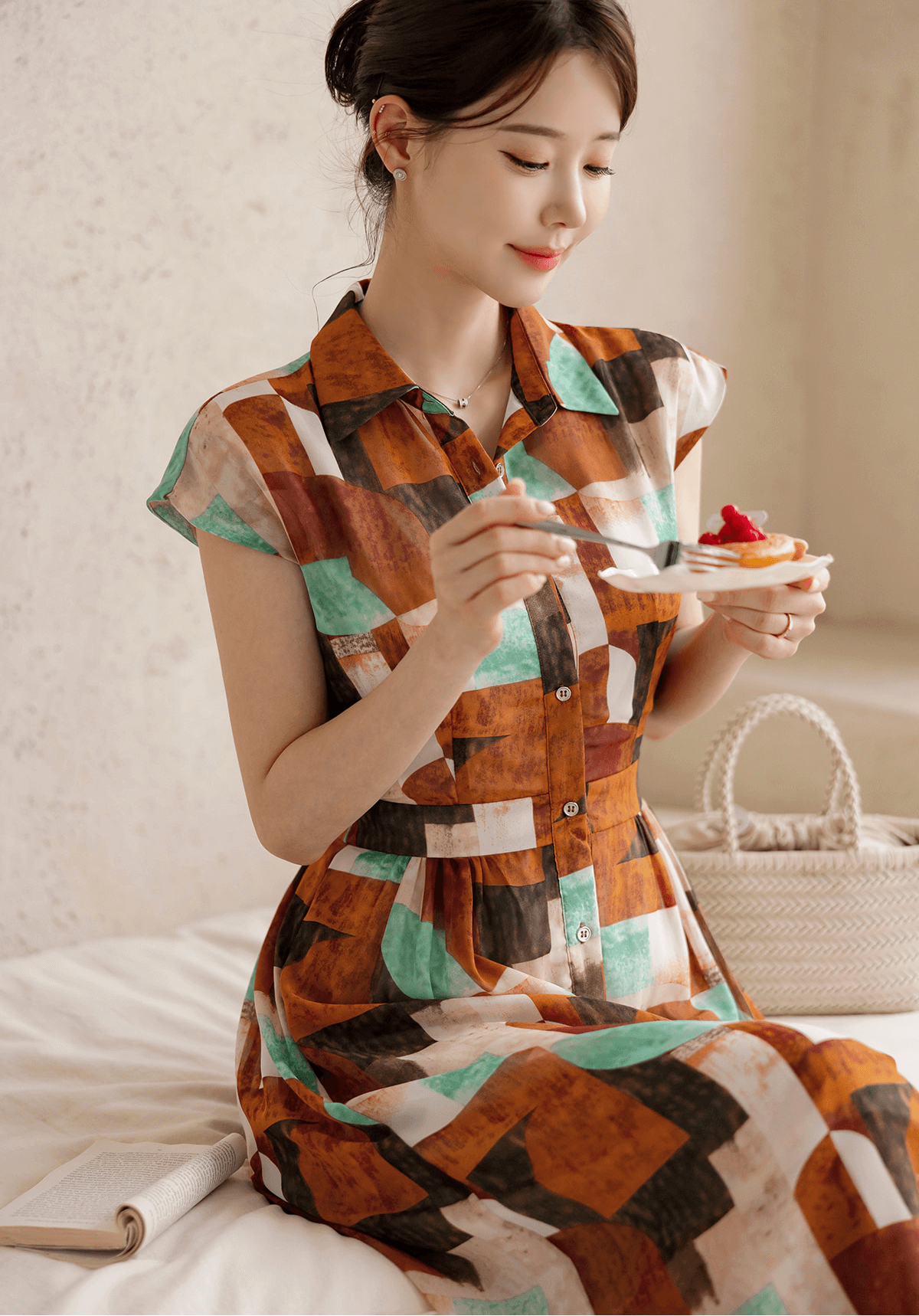 Woman in a patterned shirt dress with cap sleeves and classic collar, sitting on a bed while enjoying a dessert, with a basket handbag.
