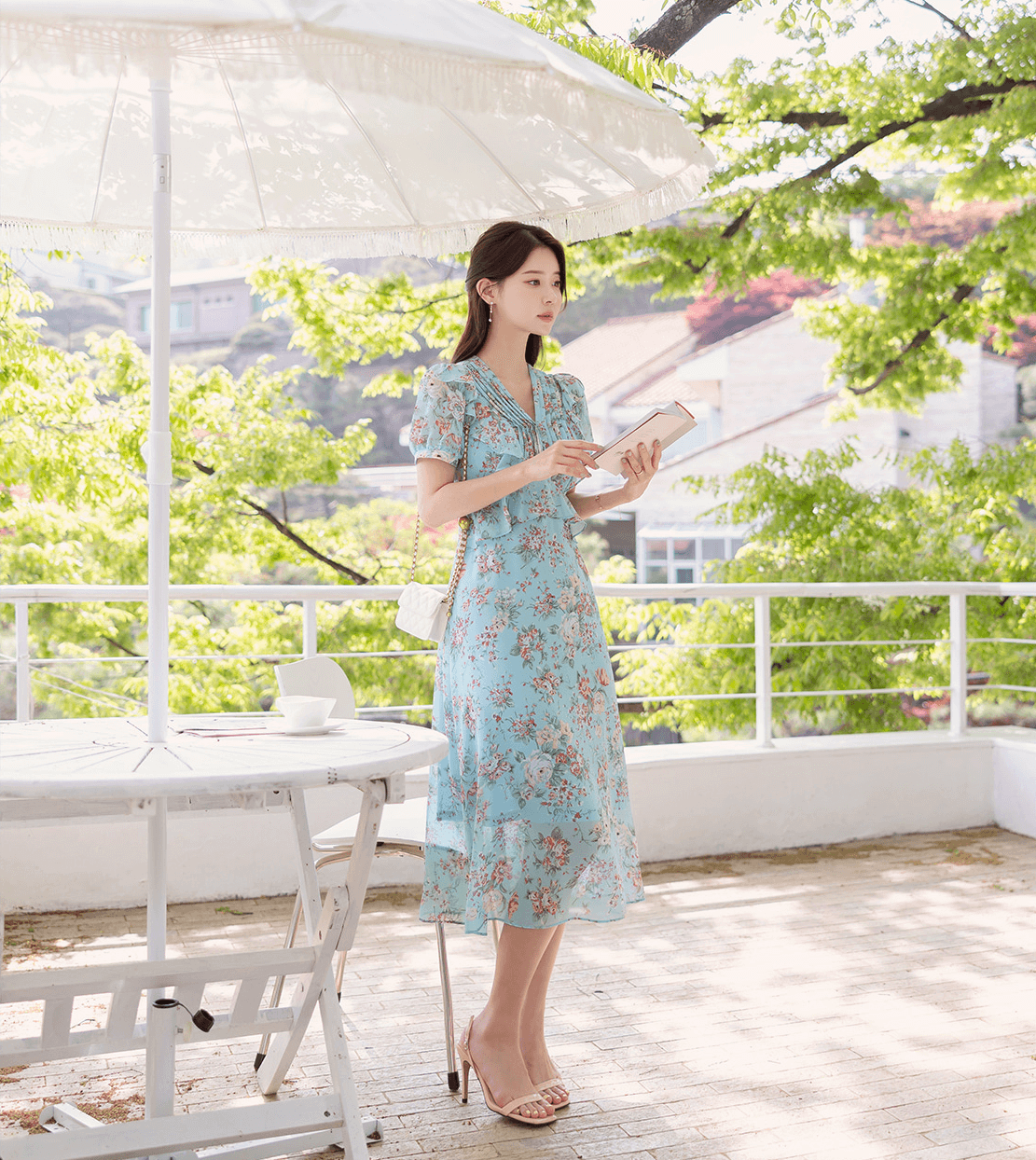 Woman wearing floral chiffon dress with pintuck bodice and peplum detail, standing outdoors with vibrant trees and white parasol.