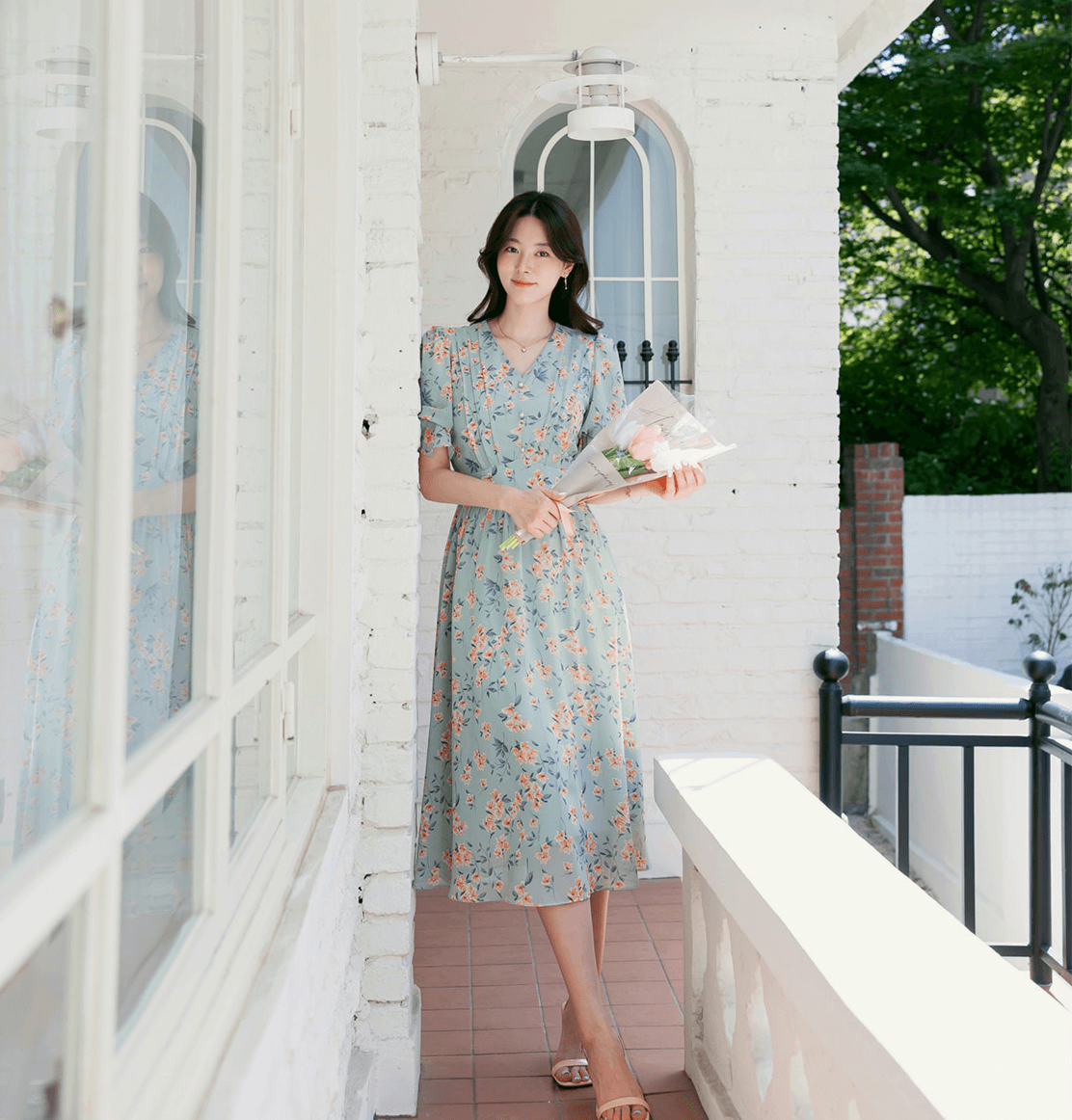 Woman wearing V-neck puff sleeve floral chiffon dress in breezy blue, holding flowers on a porch.