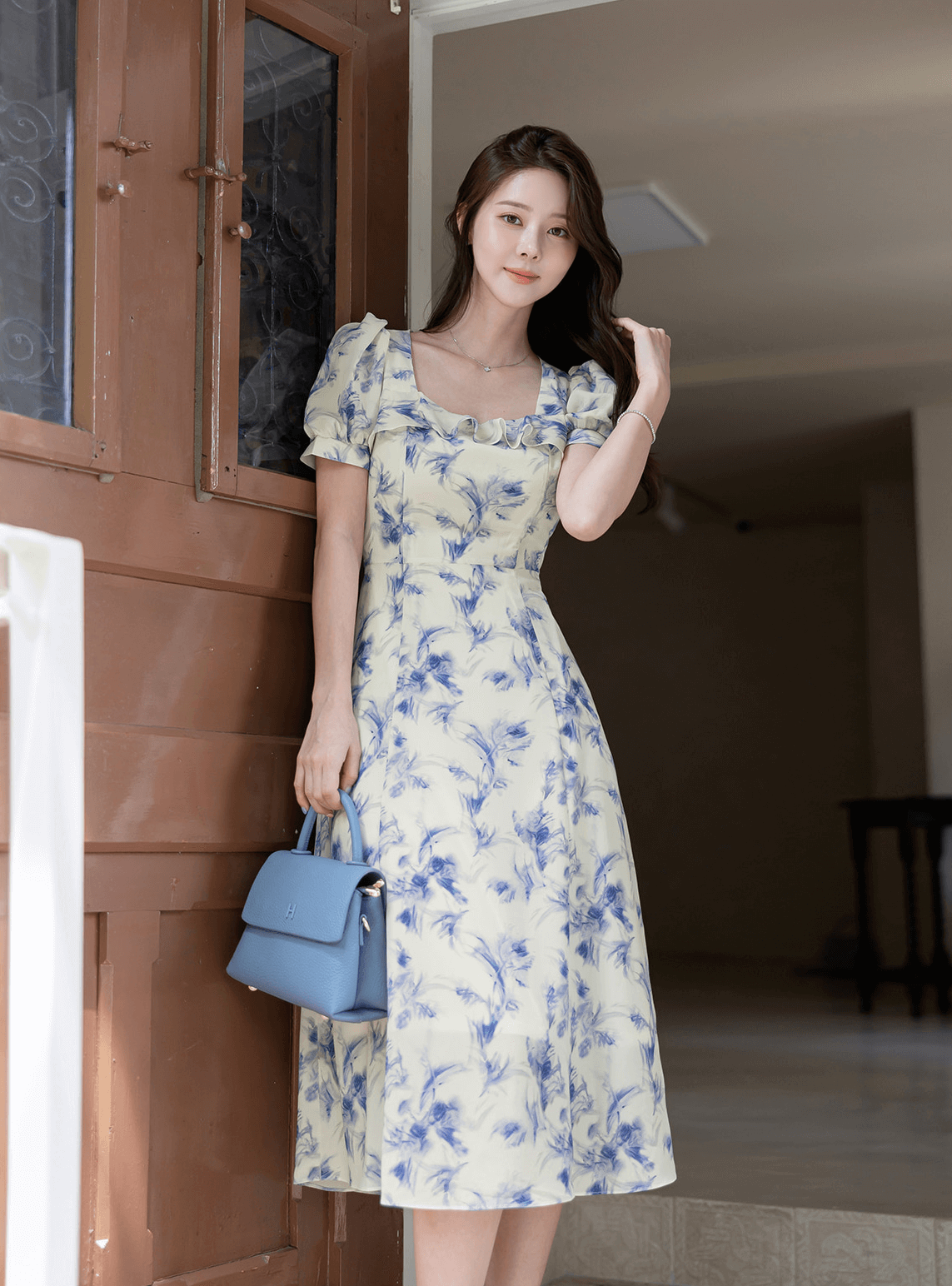 Woman wearing an elegant chiffon dress with a blue-violet abstract print, featuring a square ruffle neckline, holding a blue handbag.