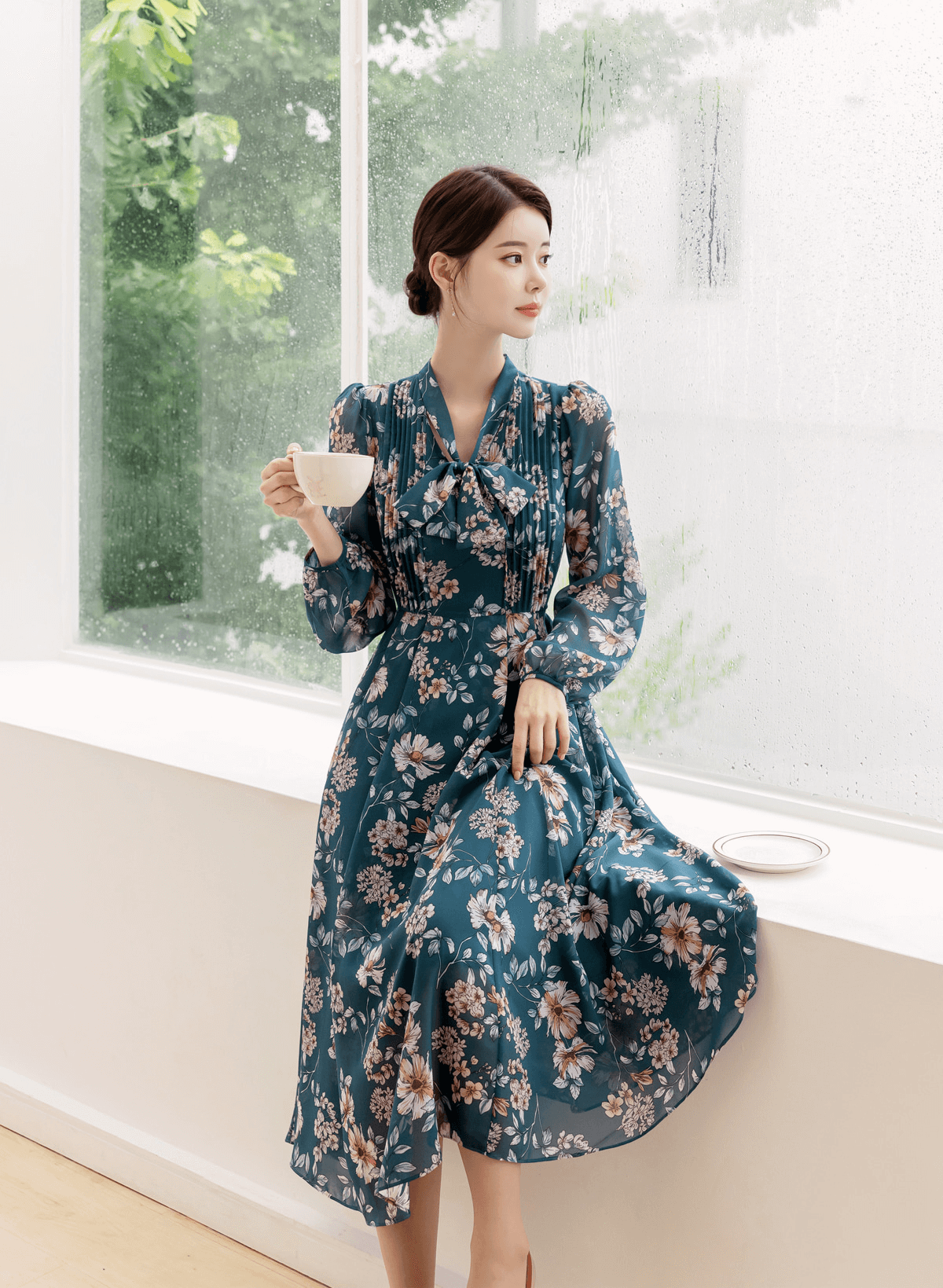 Woman in floral chiffon dress with convertible neck tie sits by window holding a cup, showcasing elegant V-neck design and flare skirt.