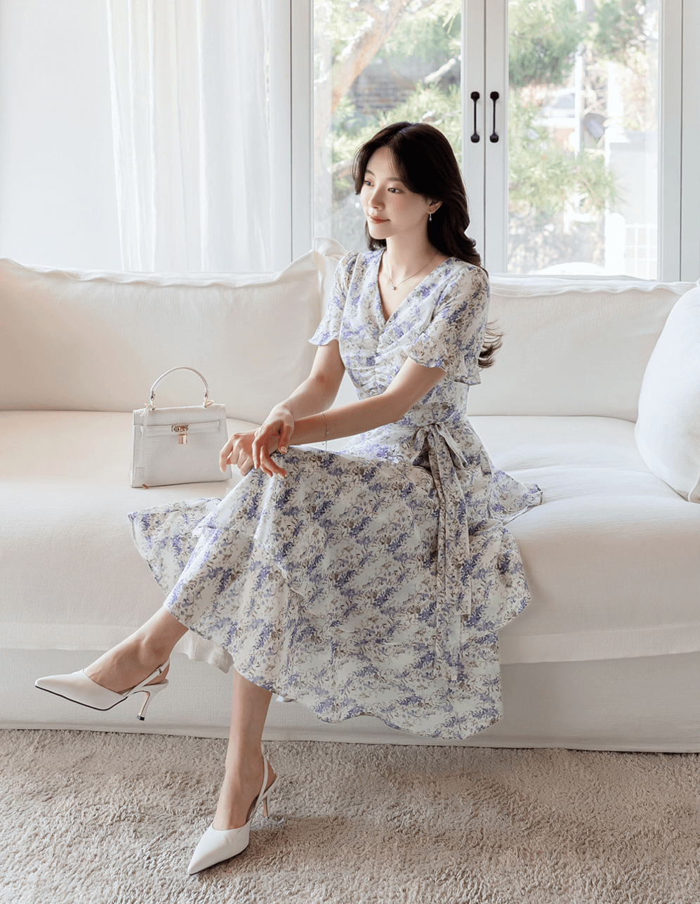 Woman wearing asymmetrical tiered floral chiffon dress with purple blossoms, sitting on a white couch, holding a handbag.
