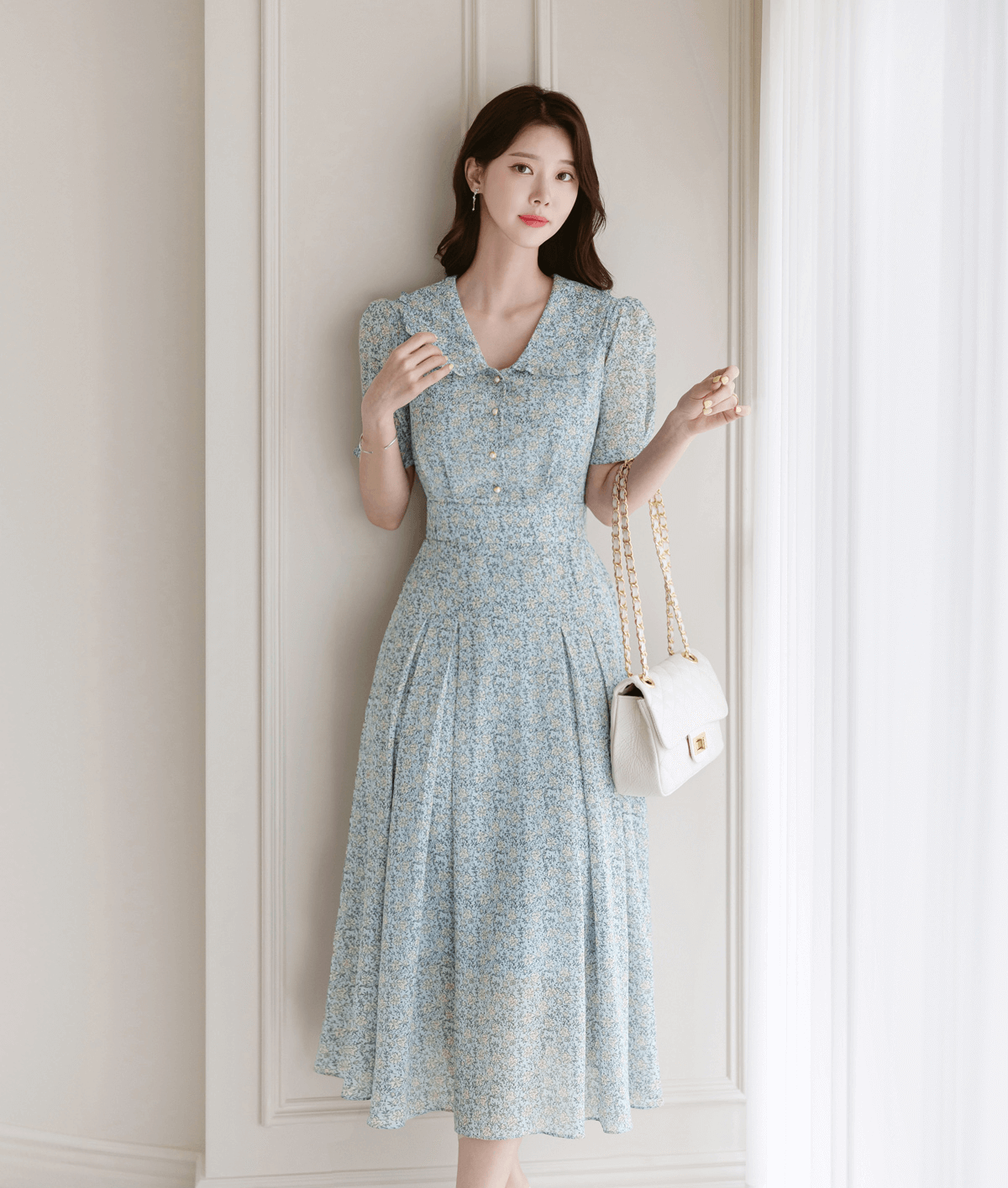 Woman in romantic floral chiffon dress with frill collar, short sleeves, and waist ribbon, standing indoors holding a white handbag.