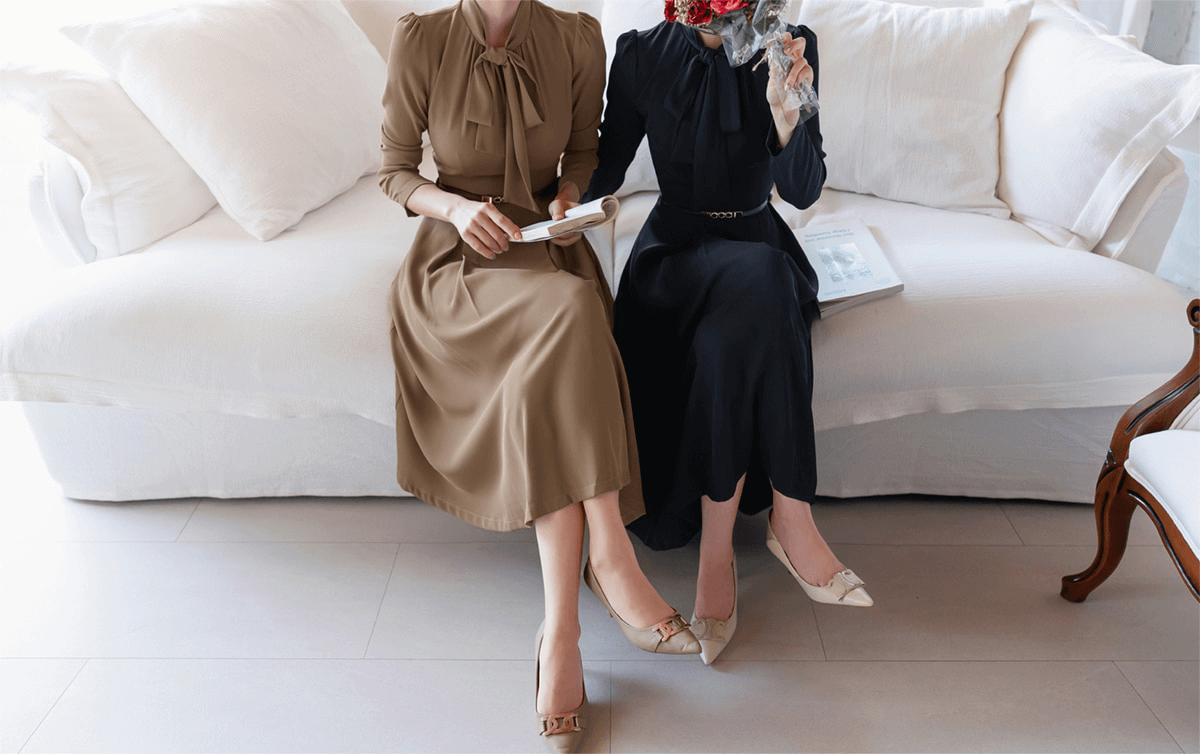 Two women sitting on a sofa wearing elegant ribbon neck pleated flare dresses in navy and beige, showcasing a timeless and classic style.