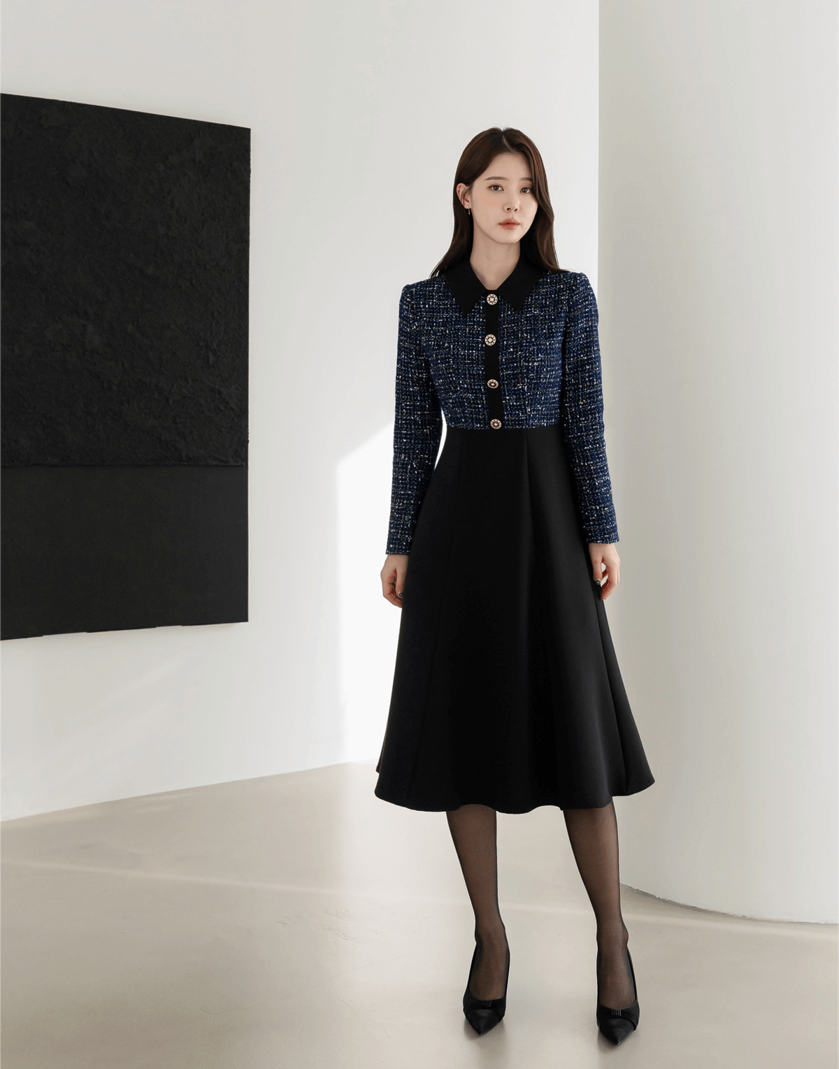Woman wearing a navy tweed and black flare dress with gold button detail, standing in a modern interior with a monochrome art piece.