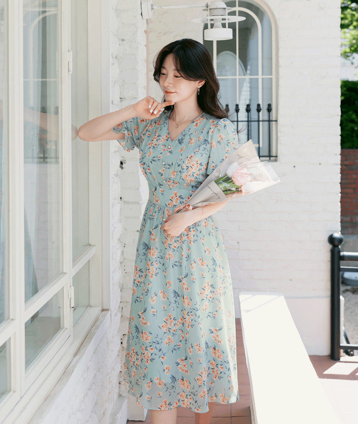 Woman wearing a V-neck puff sleeve floral chiffon dress in breezy blue, holding flowers, standing by a window.