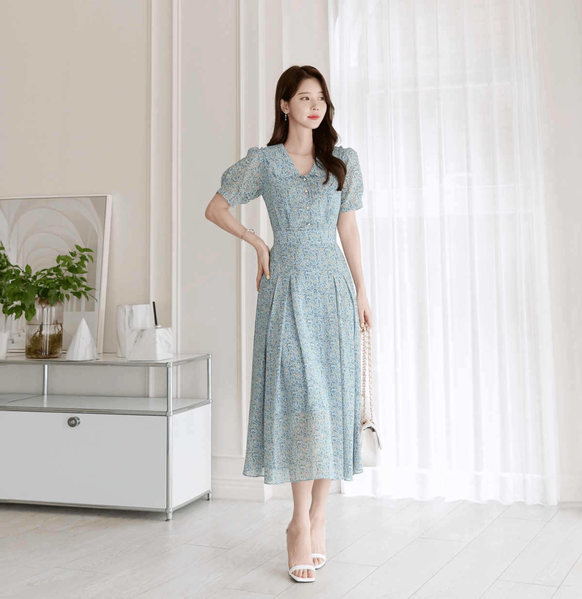Woman wearing romantic floral chiffon dress with frill collar and waist ribbon, standing elegantly in a bright room.