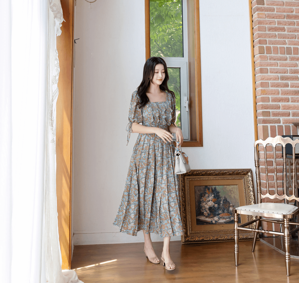 Woman wearing a pleated dress with square neckline and slit sleeves, standing in a room with wooden floor and framed artwork.