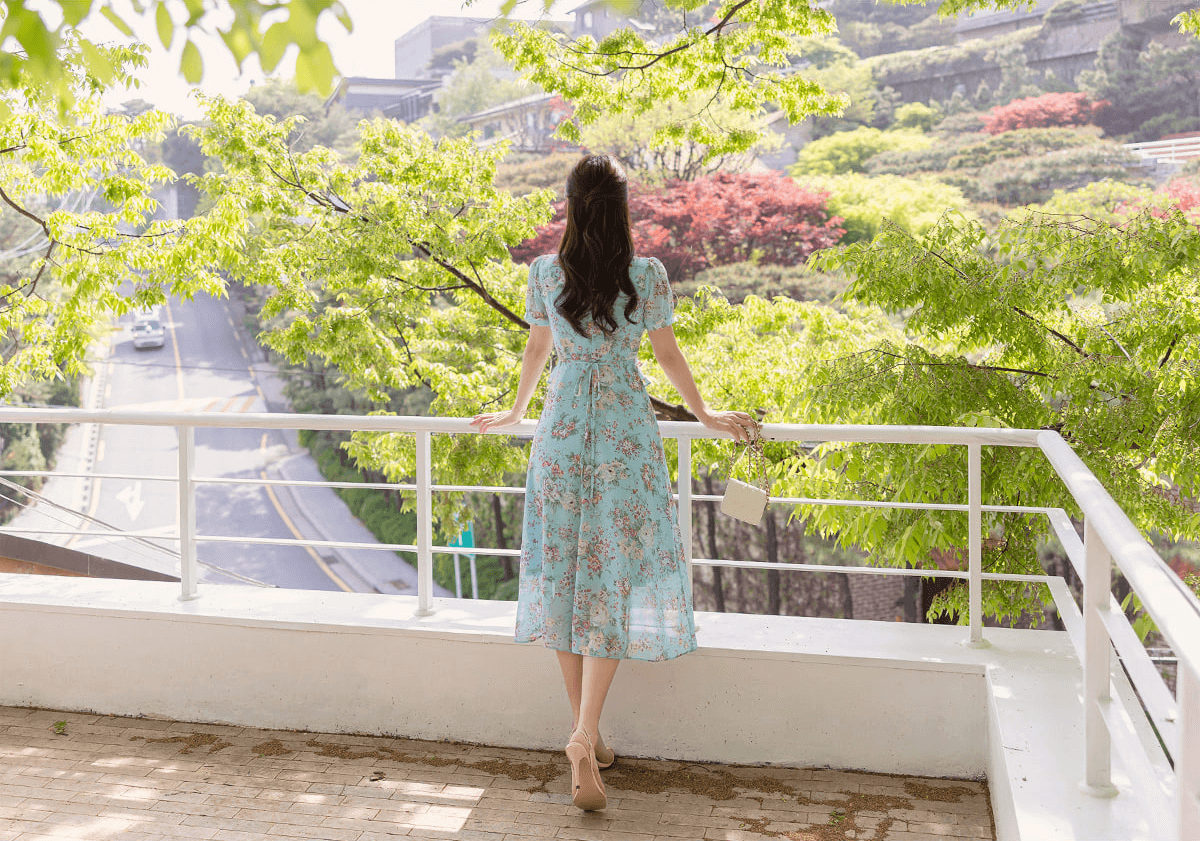 Woman wearing a floral chiffon dress with pintuck bodice and peplum detail on a balcony overlooking a scenic view.