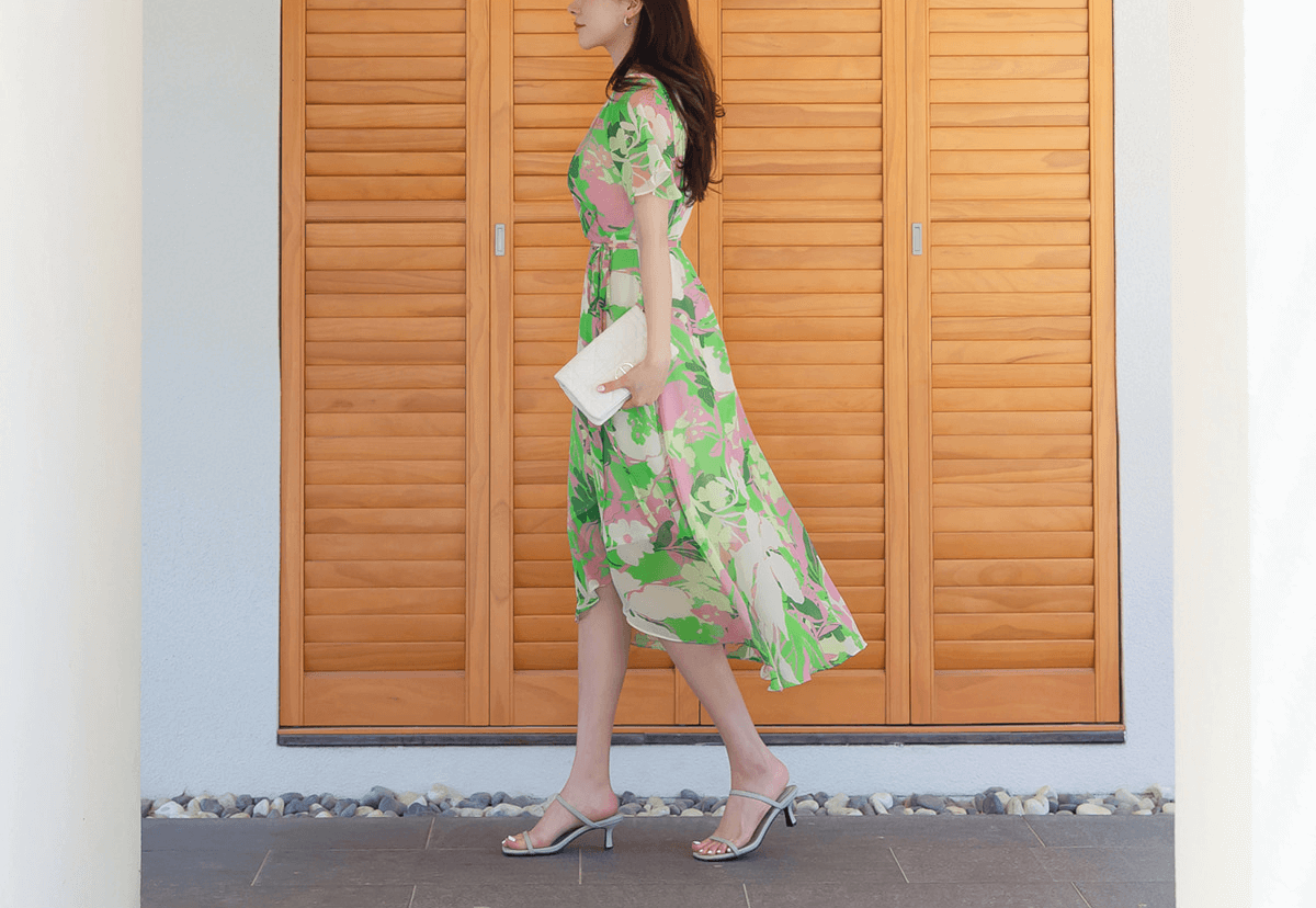 Woman in green chiffon wrap dress with tulip hem and floral print, holding a clutch, walking near wooden door.