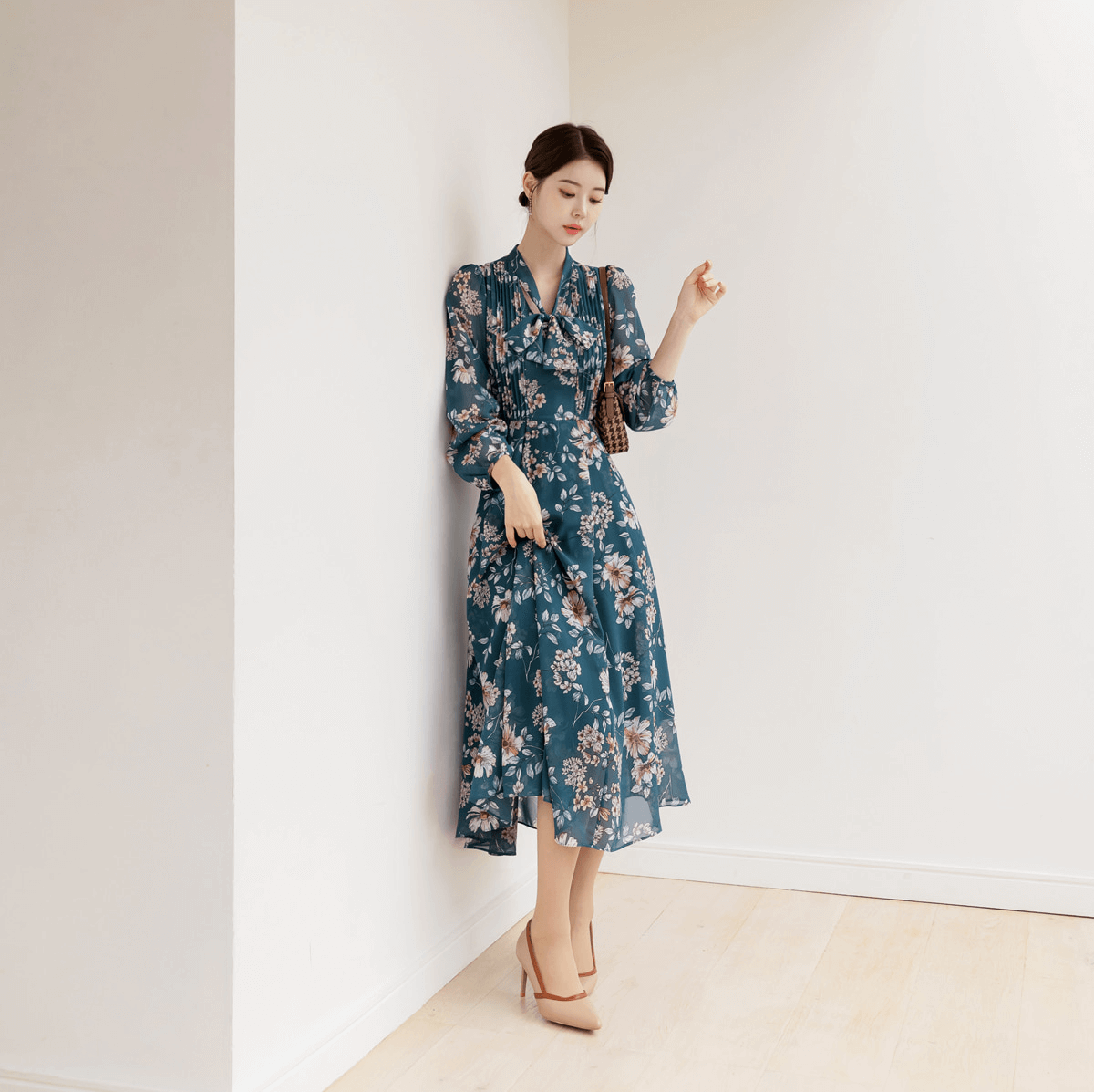 Woman in floral chiffon flare dress with V-neck and neck tie, styled elegantly, standing in minimalist room.