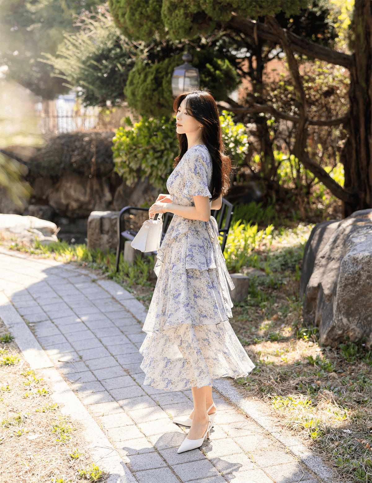 Woman wearing asymmetrical tiered floral chiffon dress with purple blossoms, standing outdoors on a sunny path.