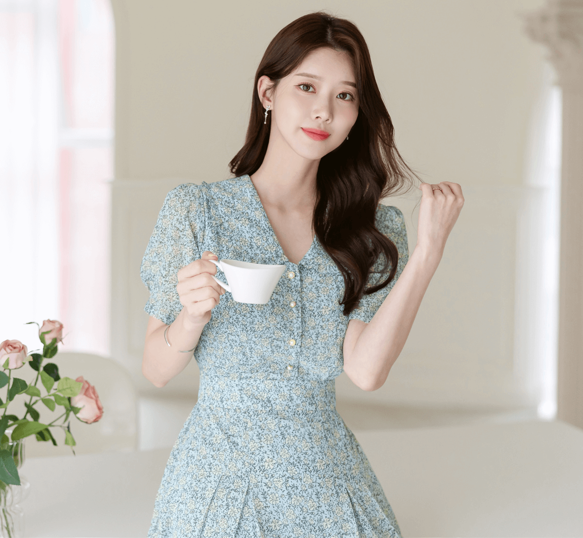 Woman in romantic floral chiffon dress with frill collar, holding a teacup in a bright room with roses in the background.