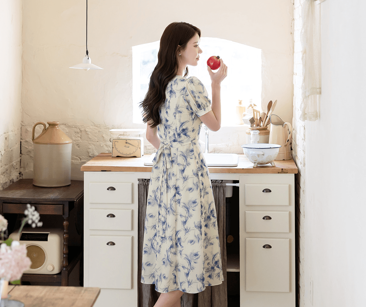 Woman in elegant chiffon dress with blue-violet abstract print standing in kitchen holding an apple.