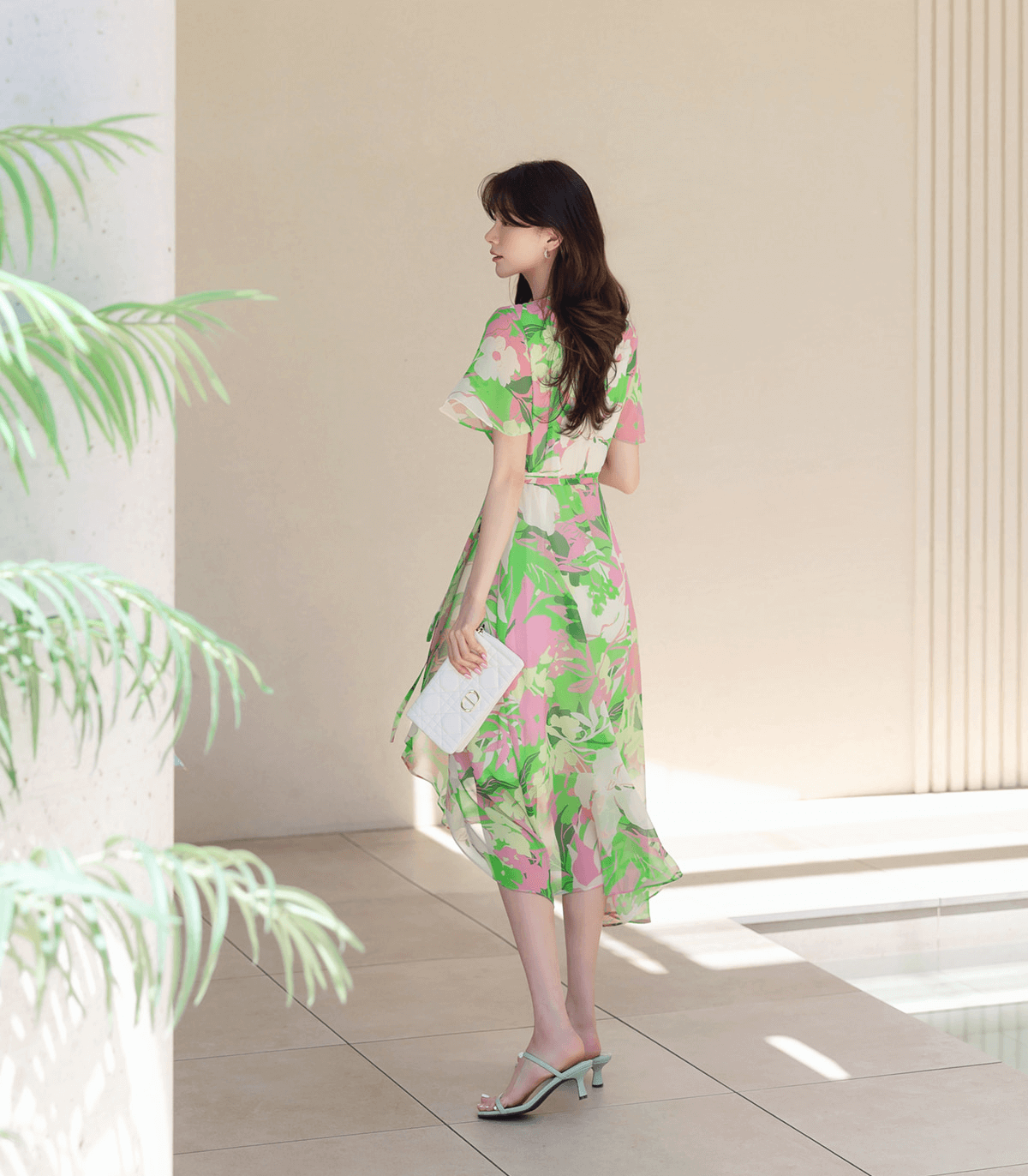 Woman in a green floral chiffon wrap dress with a tulip hem, showcasing a feminine silhouette in a sunny indoor setting.