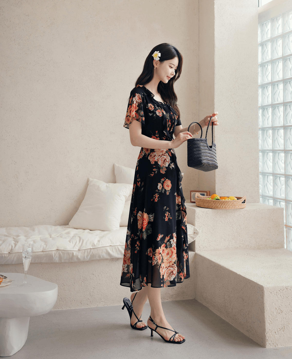 Woman in floral chiffon dress with square neckline and sheer sleeves, holds a bag, standing in a stylish room.
