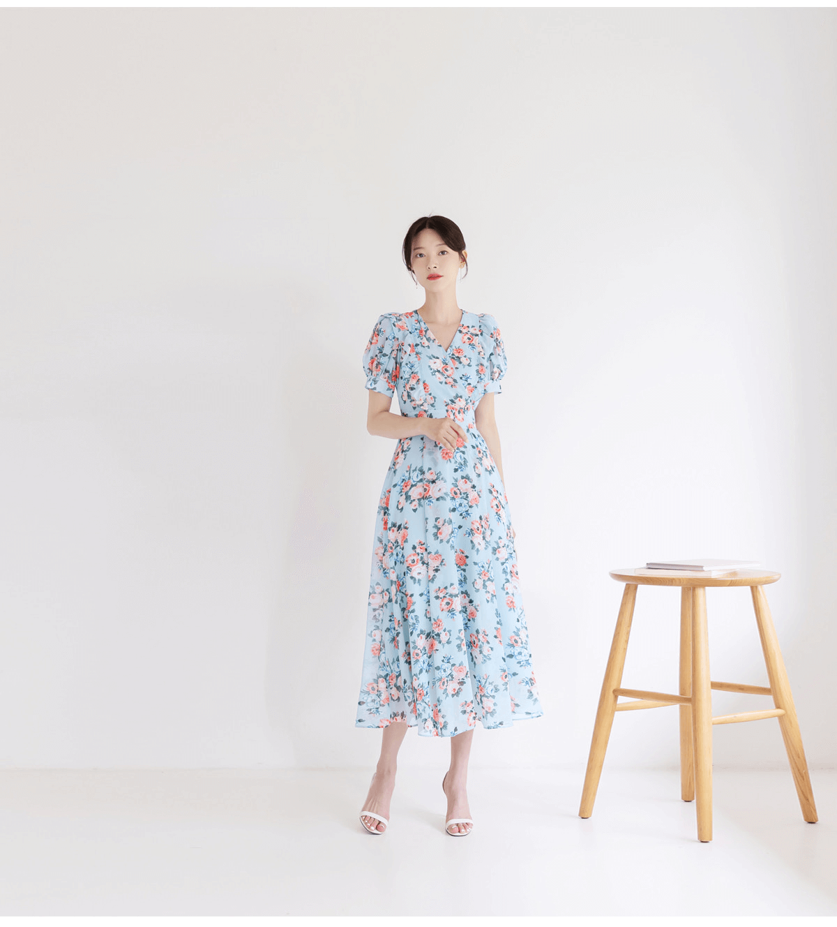 Woman wearing blue floral chiffon dress with V-neck and puff sleeves, standing next to stool in minimalistic room.