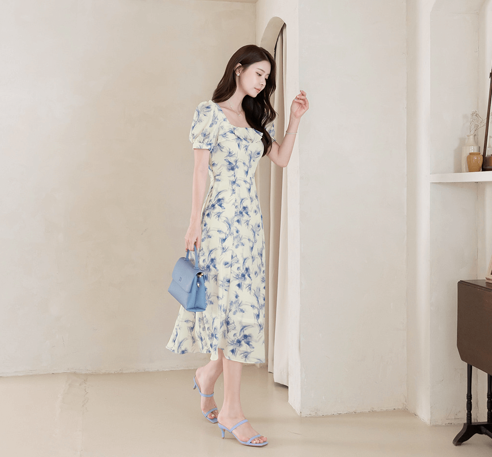 Woman in elegant chiffon dress with blue-violet abstract print and square ruffle neckline, holding a matching blue handbag.