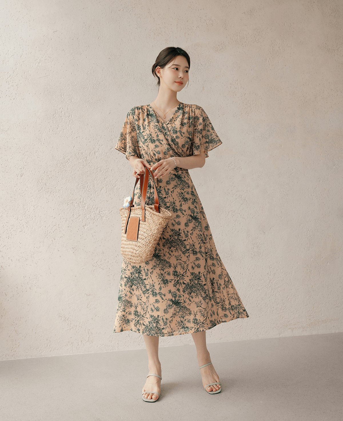 Woman wearing a goddess-inspired wrap dress with graceful wing sleeves, holding a straw bag, against a neutral wall.