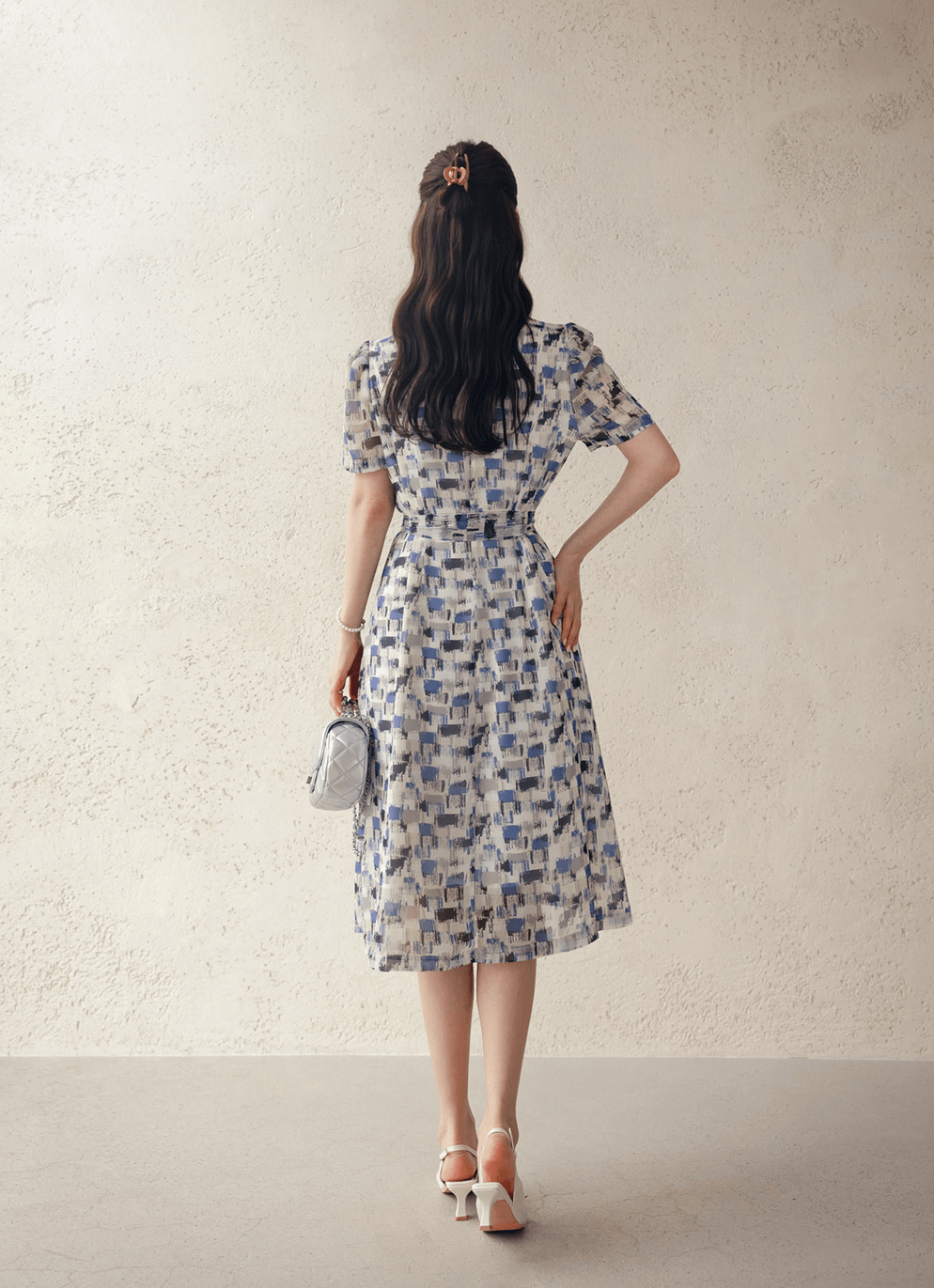 Back view of woman wearing breezy chiffon shirt dress with abstract blue pattern and belt, holding a small purse, in a light room