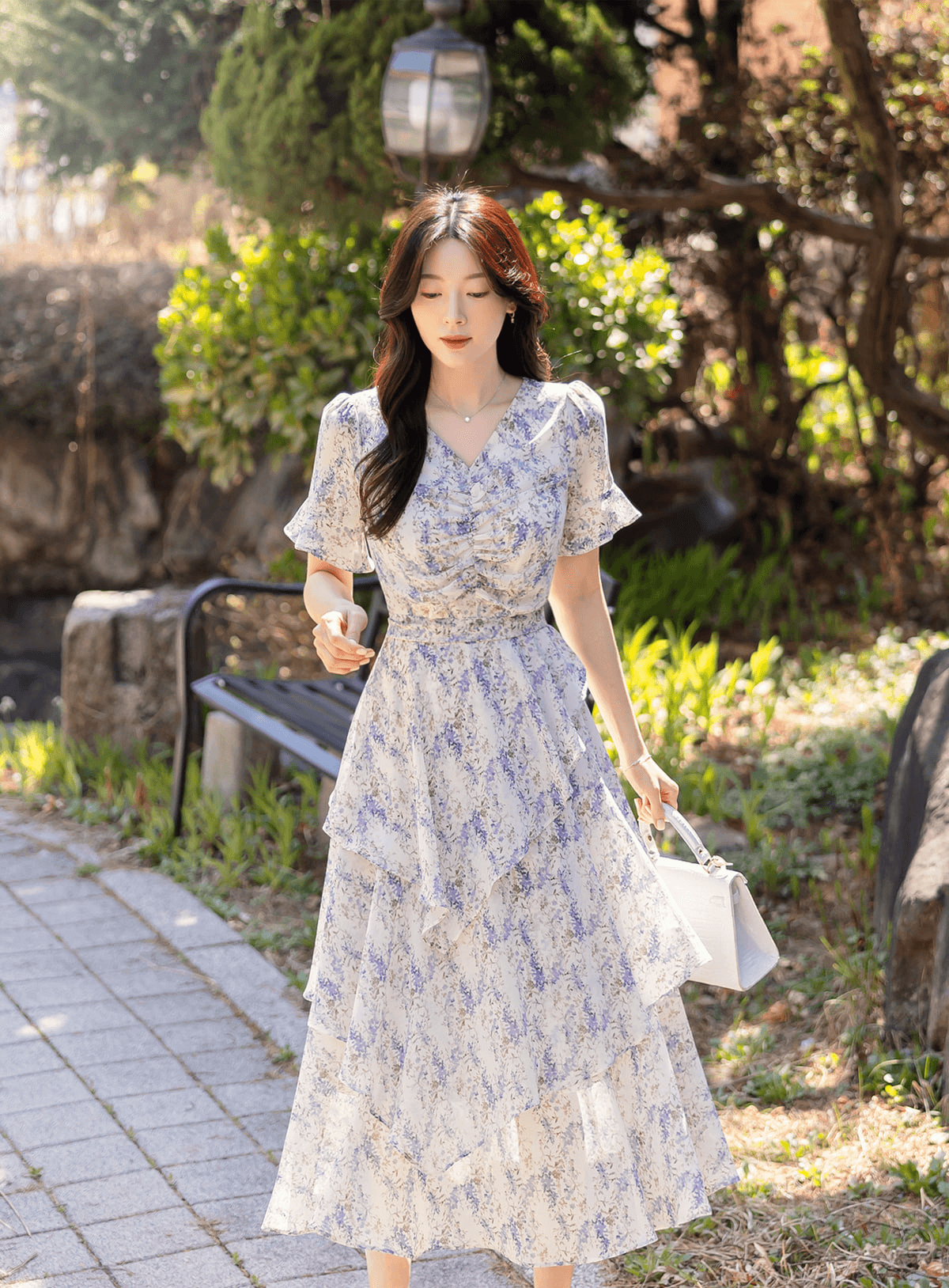 Woman wearing an asymmetrical tiered floral chiffon dress with purple blossoms, featuring a V-neckline and ruffled skirt on a sunny path.