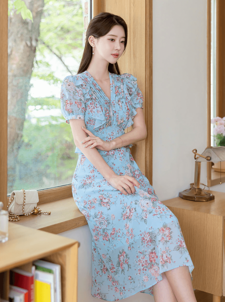 Light blue floral chiffon dress with pintuck bodice, peplum waist, and puffed sleeves worn by woman sitting by window.