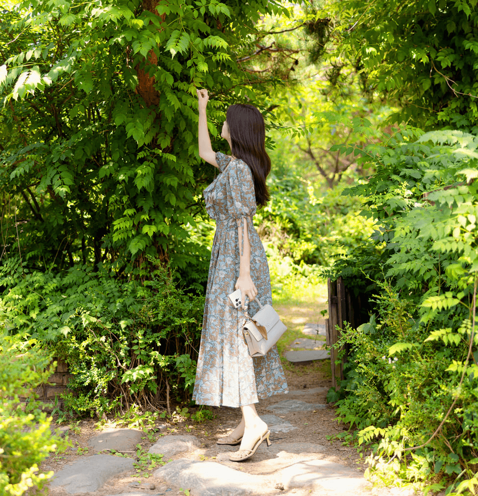 Woman in floral pleated dress with slit sleeves and ribbon ties, walking in a vibrant green garden, holding a stylish handbag.