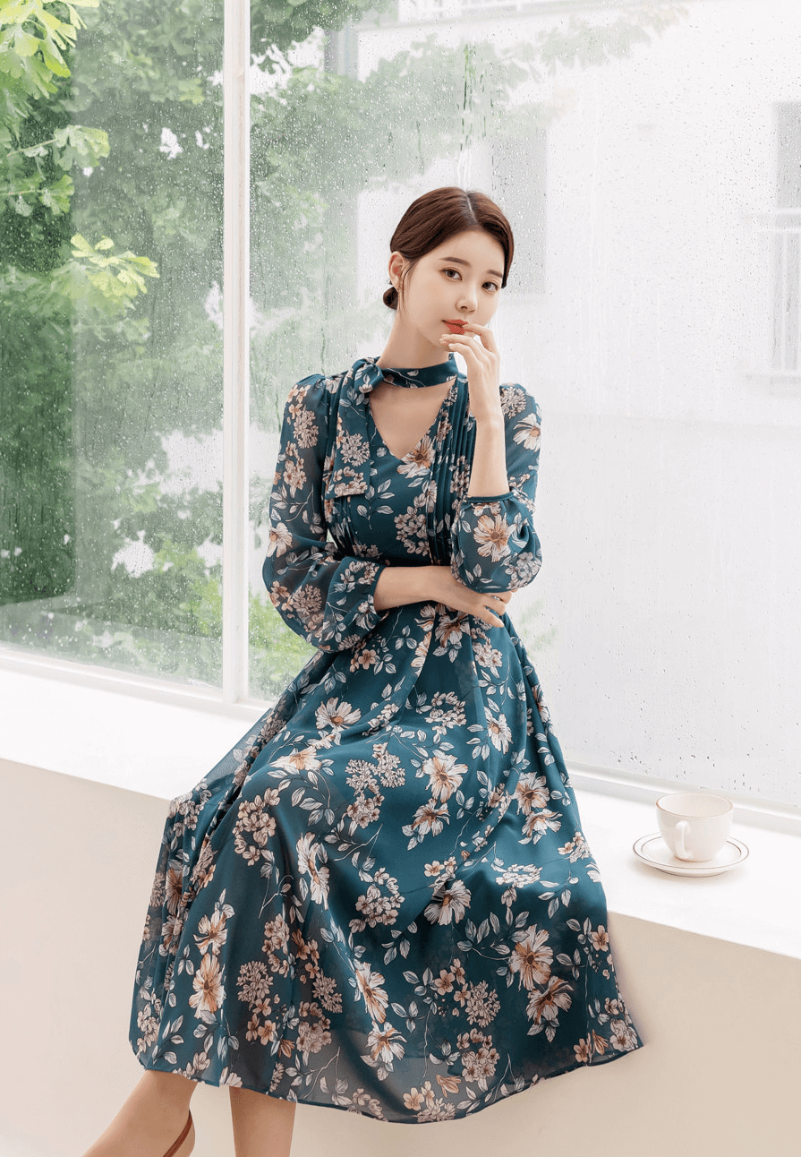 Woman wearing a floral chiffon flare dress with V-neck and convertible neck tie sitting by a window.