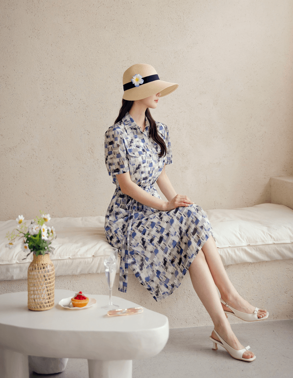Woman wearing a breezy chiffon shirt dress with abstract blue pattern and belt, seated stylishly on a bench, with summer hat and heels.