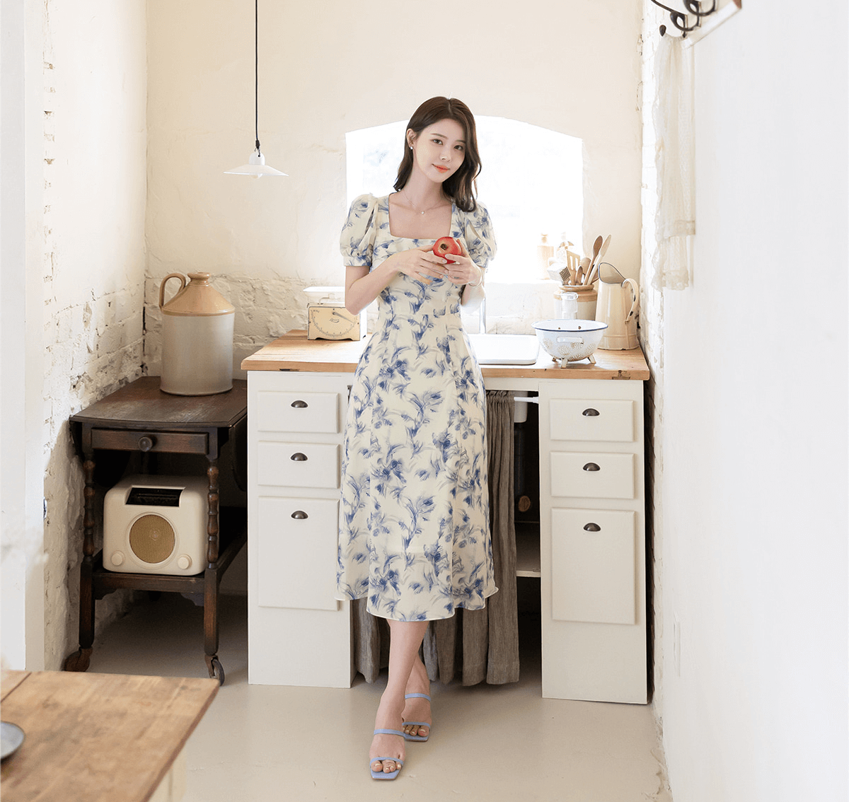 Woman in elegant chiffon dress with blue-violet print and square neckline, standing in cozy kitchen holding a red fruit.