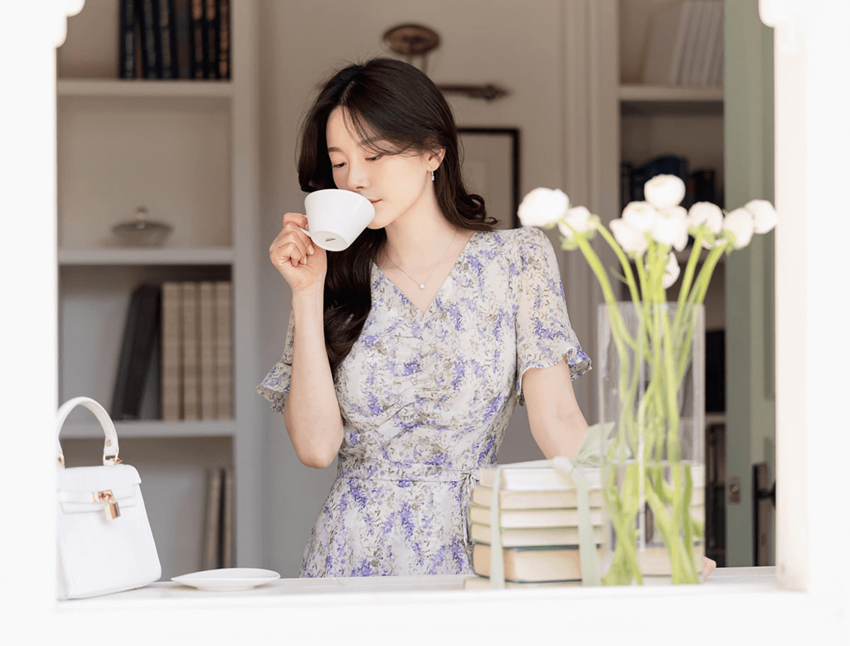 Woman in floral chiffon dress enjoying coffee at a cozy home with books and flowers in the foreground.