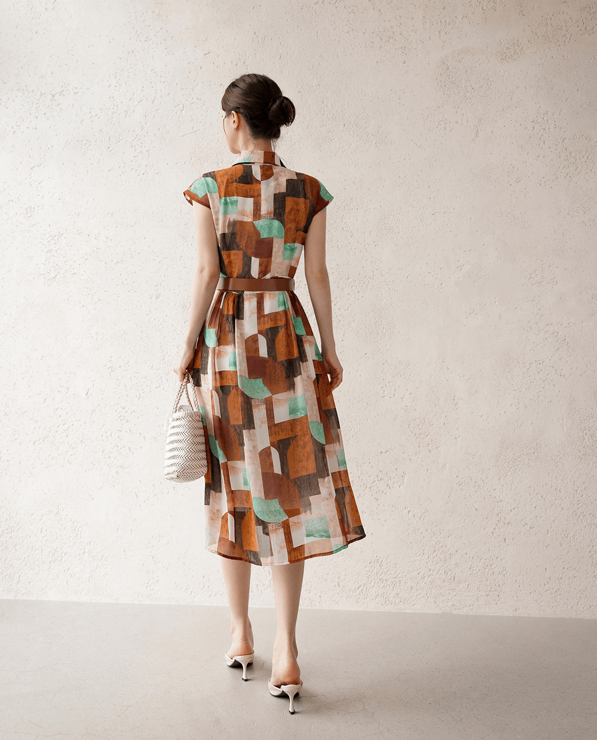 Back view of a woman wearing a patterned shirt dress with cap sleeves, classic collar, and faux leather belt, holding a woven bag.