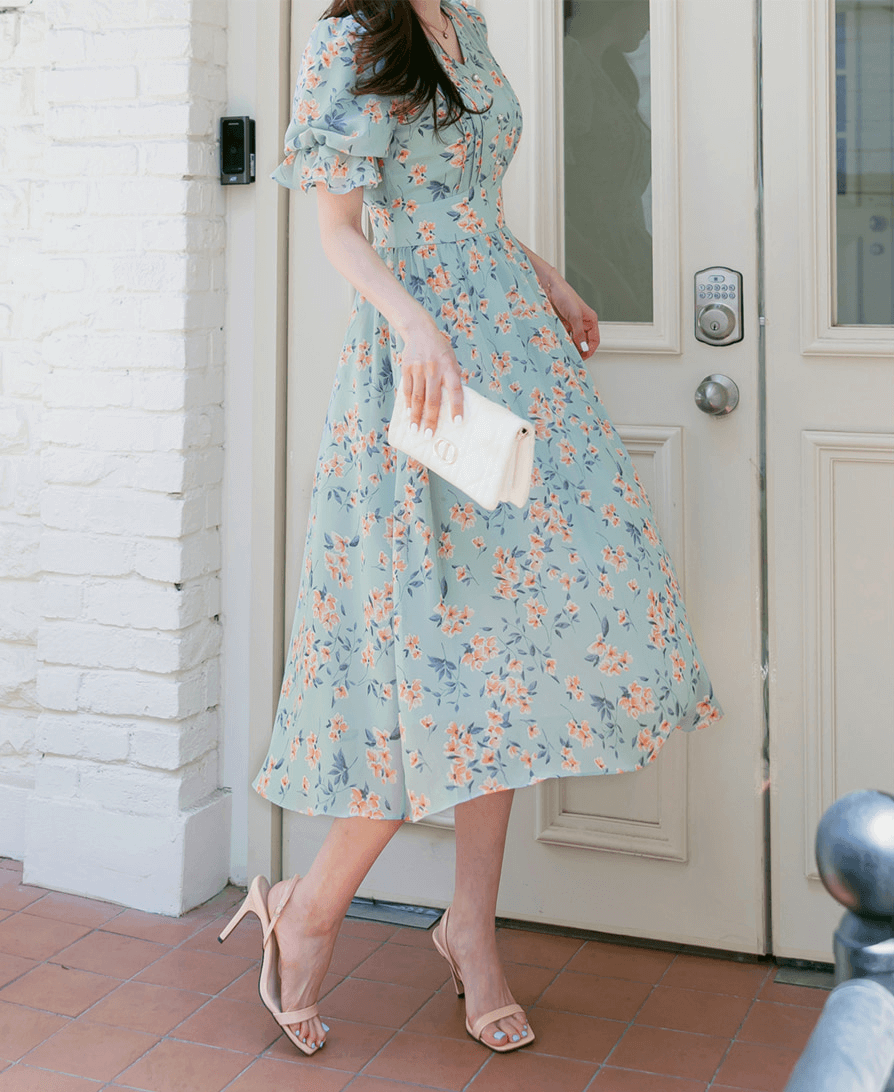 Woman wearing a V-neck puff sleeve floral chiffon dress in breezy blue, holding a clutch, and standing near a door.
