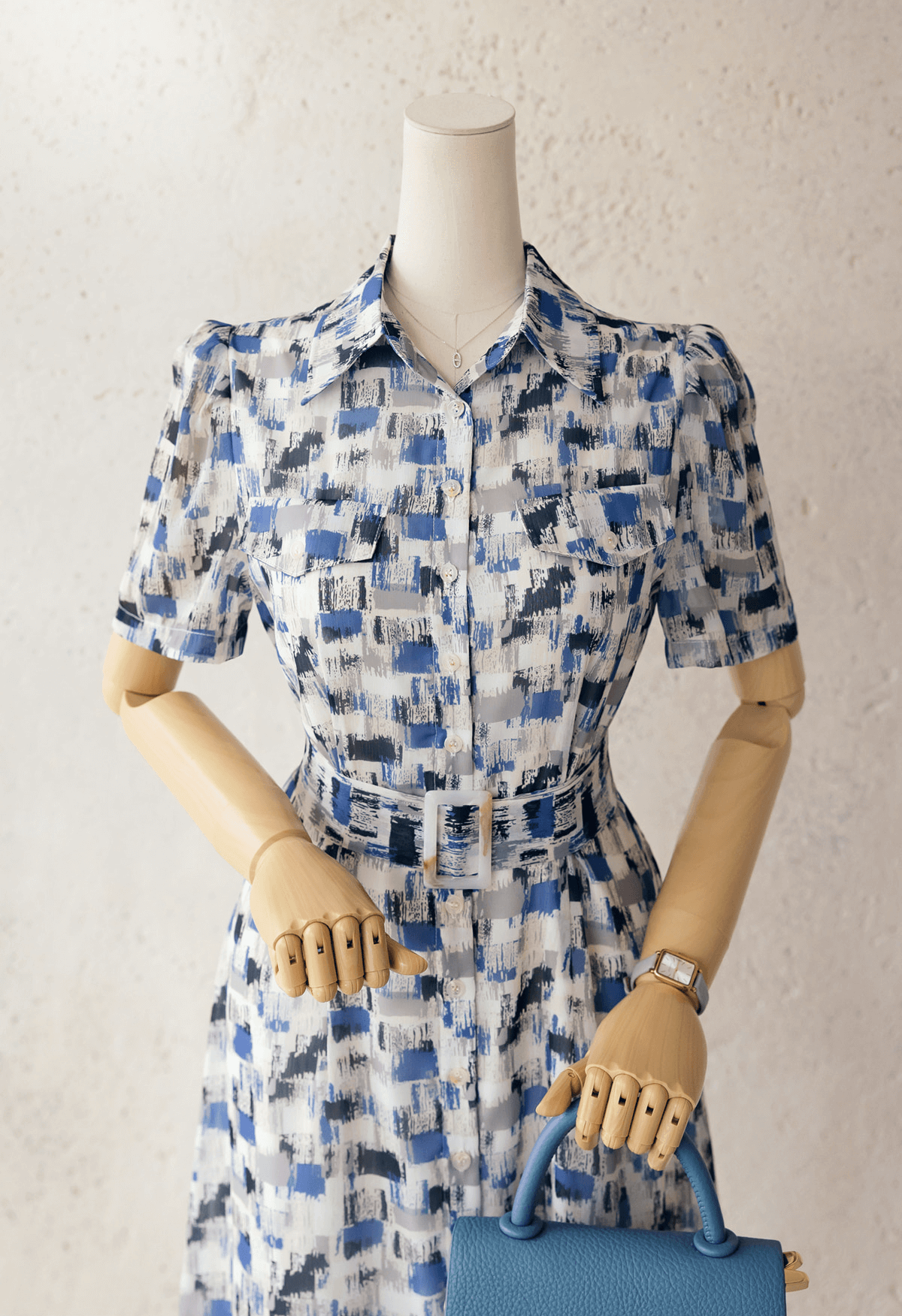 Chiffon shirt dress in abstract blue pattern with belt displayed on mannequin, featuring collar and short sleeves, holding a blue handbag.