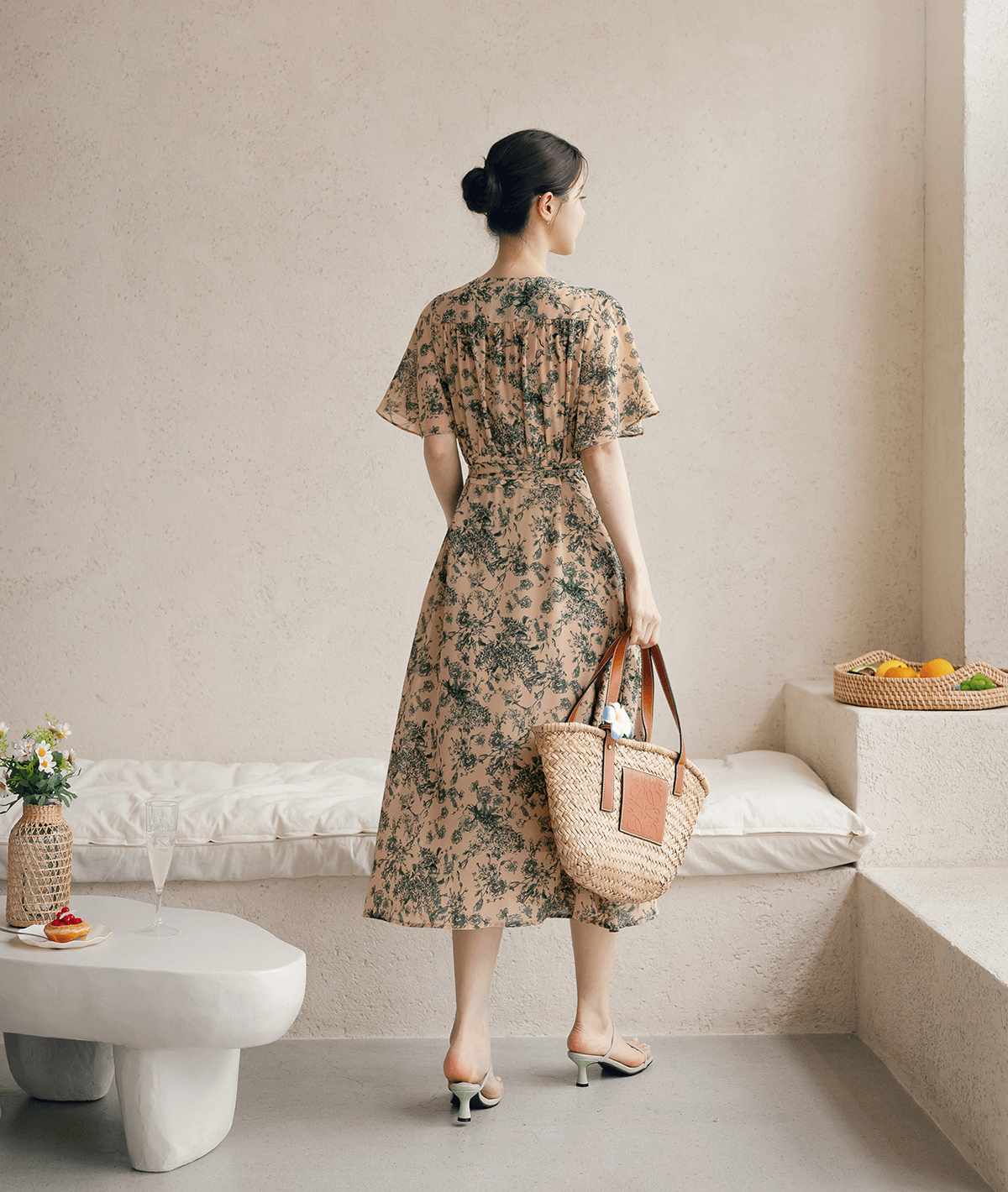 Woman in goddess-inspired wrap dress with wing sleeves, holding a straw bag, standing by a minimalistic bench with a fruit basket.
