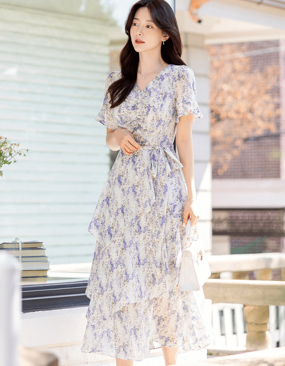Woman wearing asymmetrical tiered floral chiffon dress with purple blossoms and puffed sleeves in a sunny outdoor setting.