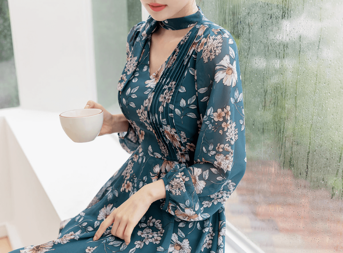 Woman wearing a floral chiffon flare dress with V-neck and neck tie, sitting by a window, holding a cup. Perfect for elegant styling.