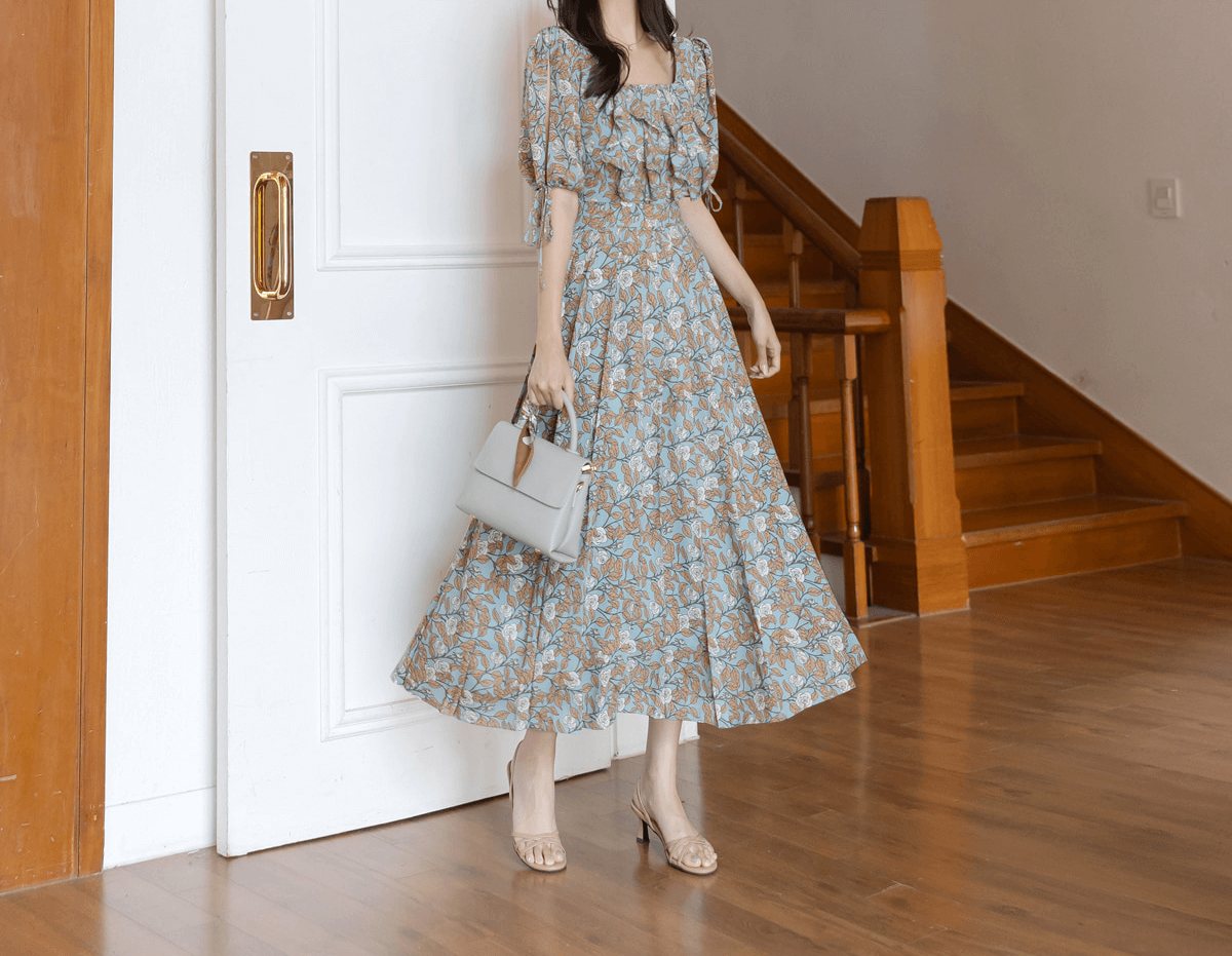 Woman wearing pleated dress with slit sleeves and ribbon ties, standing beside staircase, holding a white handbag.