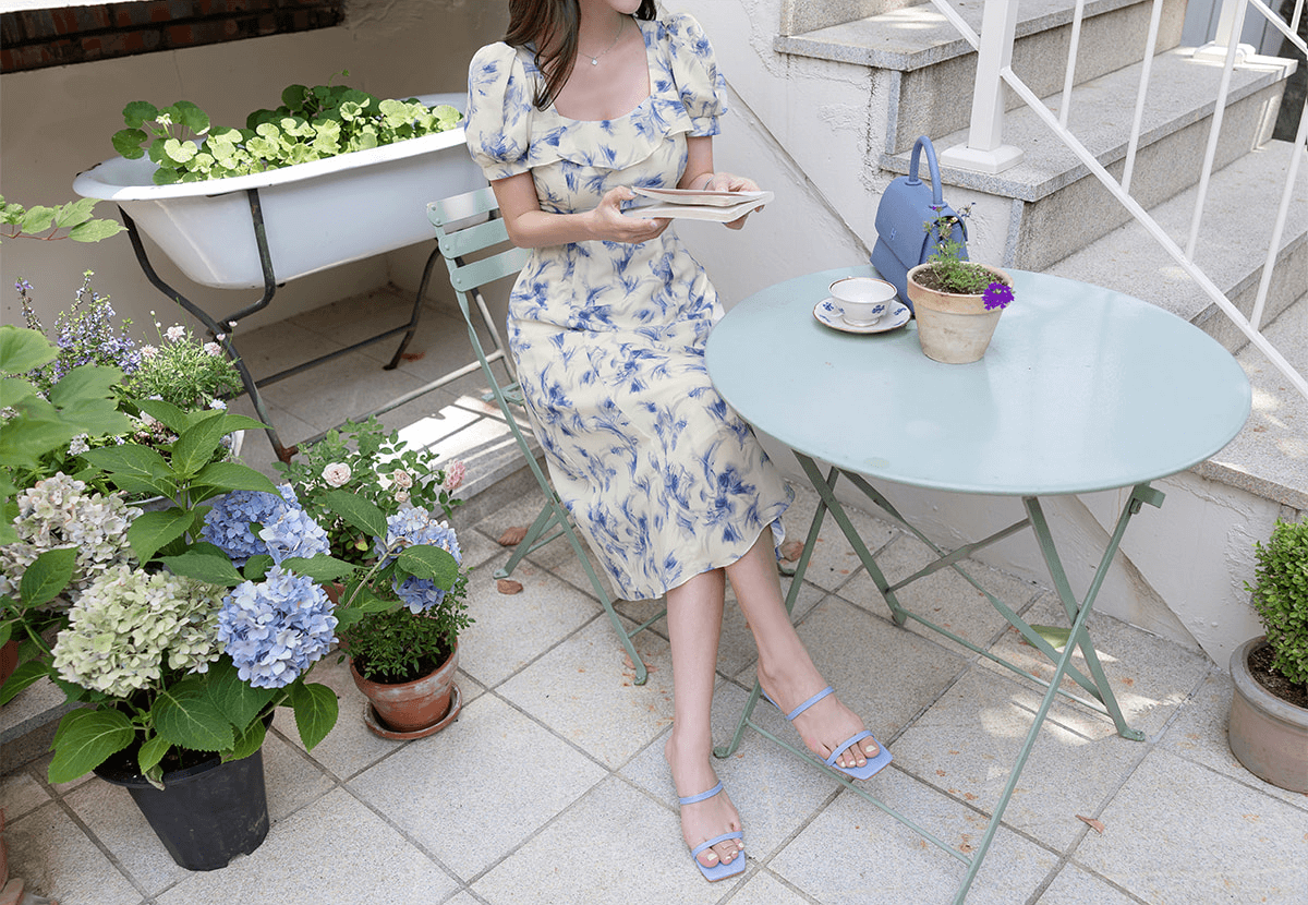 Woman in elegant chiffon dress with blue-violet abstract print enjoying a coffee at a patio table surrounded by hydrangeas.
