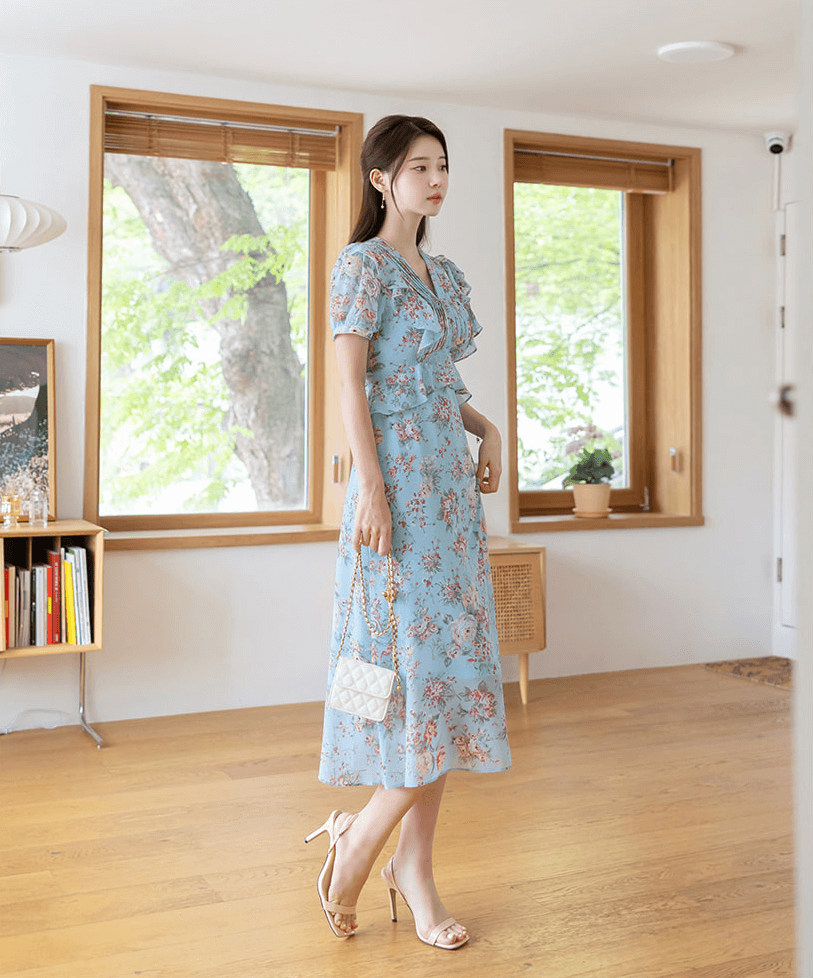 Woman in sky blue floral chiffon dress with pintuck bodice and peplum detail standing in a room with wooden floors and large windows.