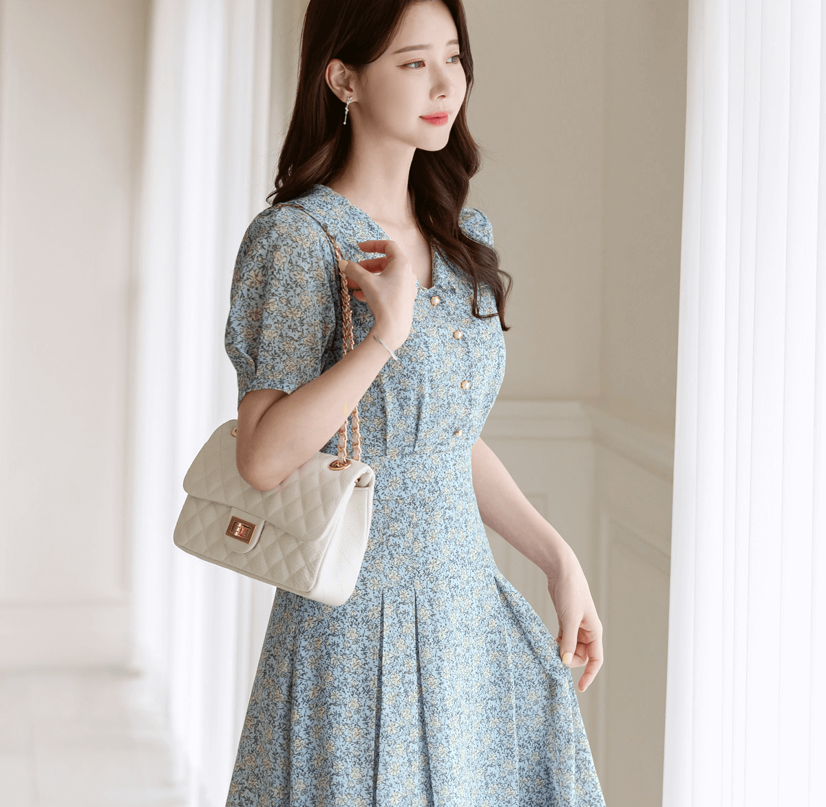 Woman in romantic floral chiffon dress with frill collar and waist ribbon, holding a white quilted purse in bright room.