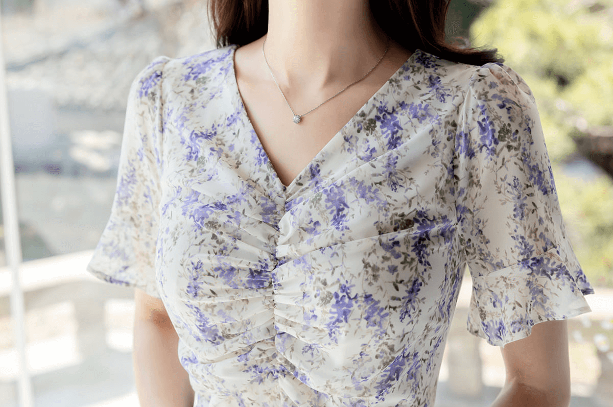 Close-up of a woman wearing a floral chiffon dress with a V-neckline and purple blossoms, showcasing soft puffed sleeves and delicate ruching.