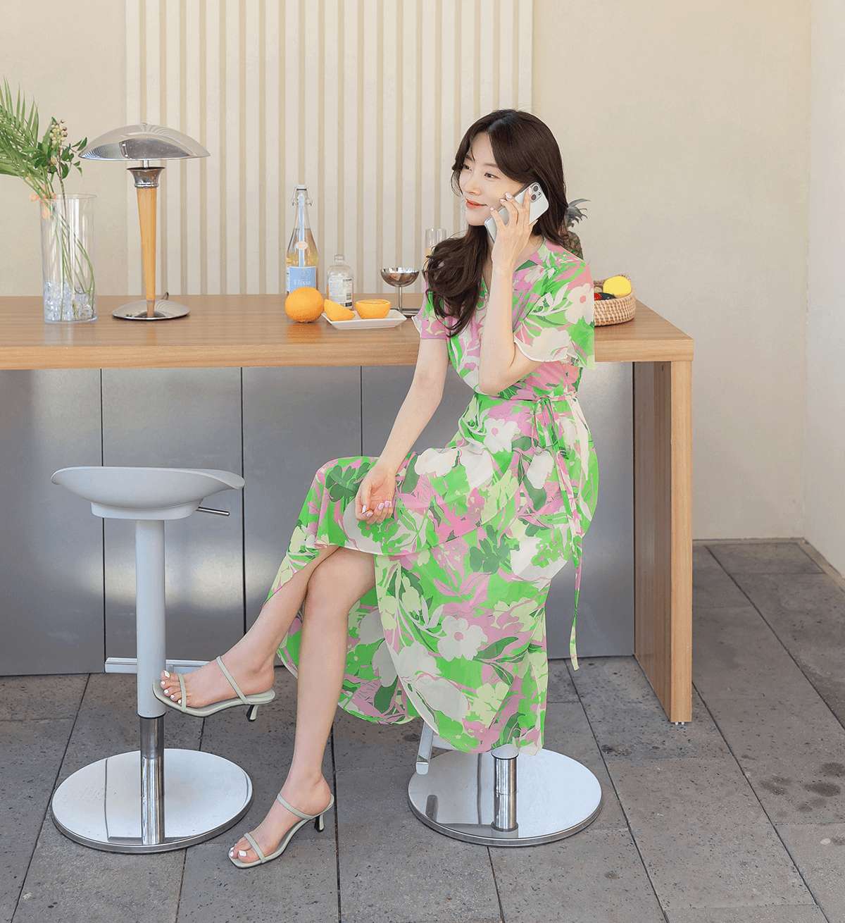 Woman in a green floral chiffon wrap dress with tulip hem sitting at a modern bar, exuding vacation style.