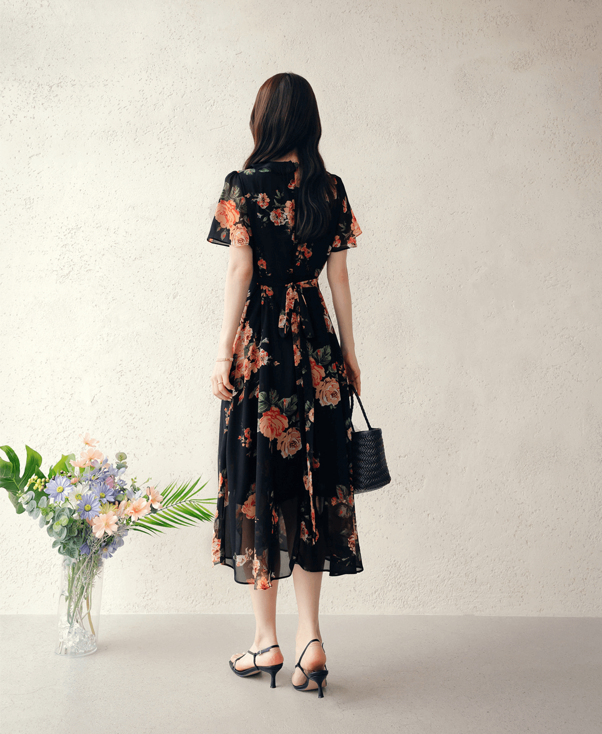 Woman wearing a floral chiffon dress with sheer wing sleeves and square neckline, carrying a black handbag, standing near flowers.