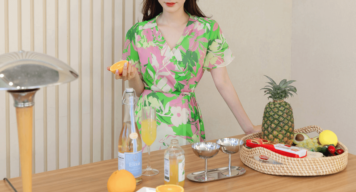 Woman in green floral chiffon wrap dress with tulip hem at kitchen counter with pineapple and orange.